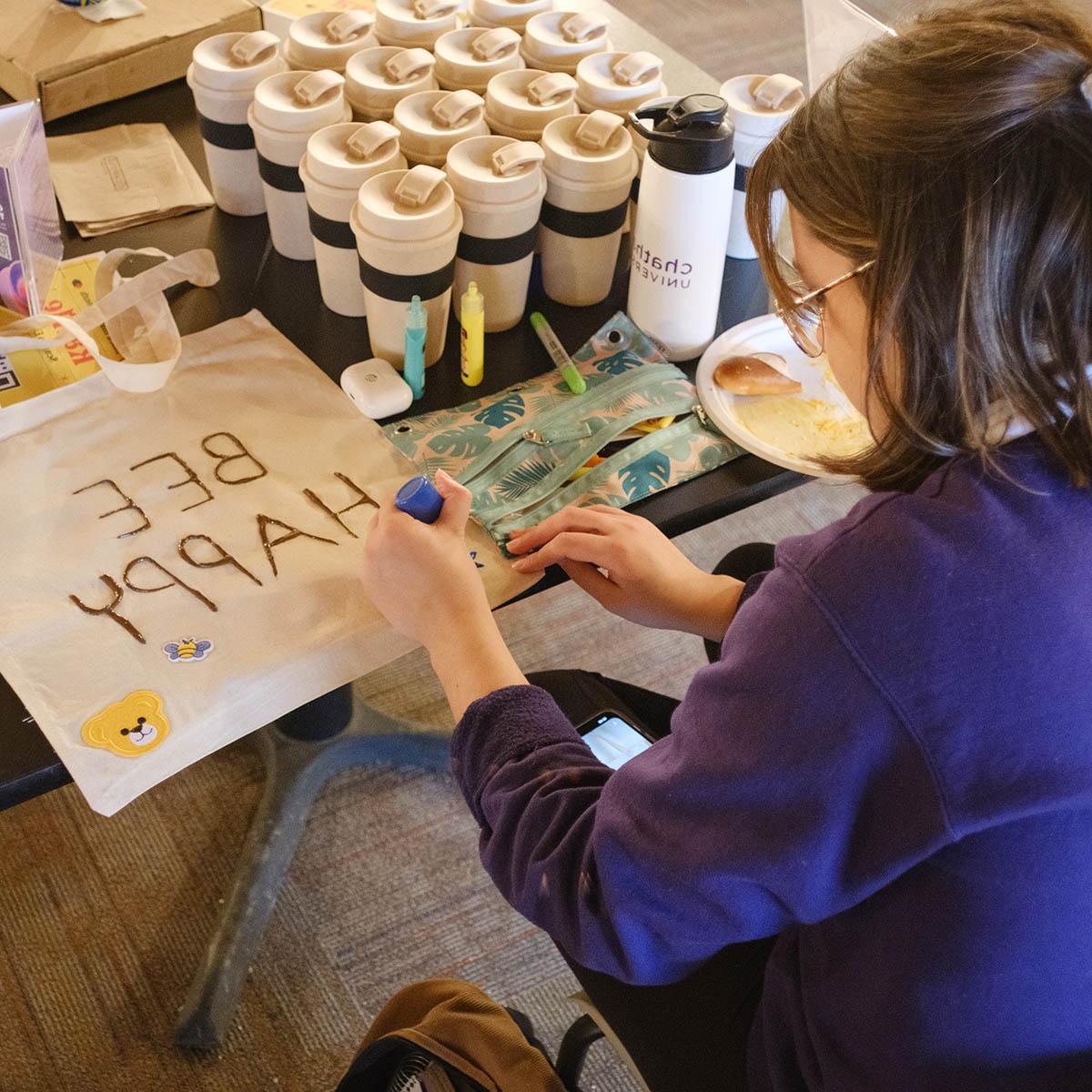 A student writes BEE HAPPY on a tote bag with puffy paint